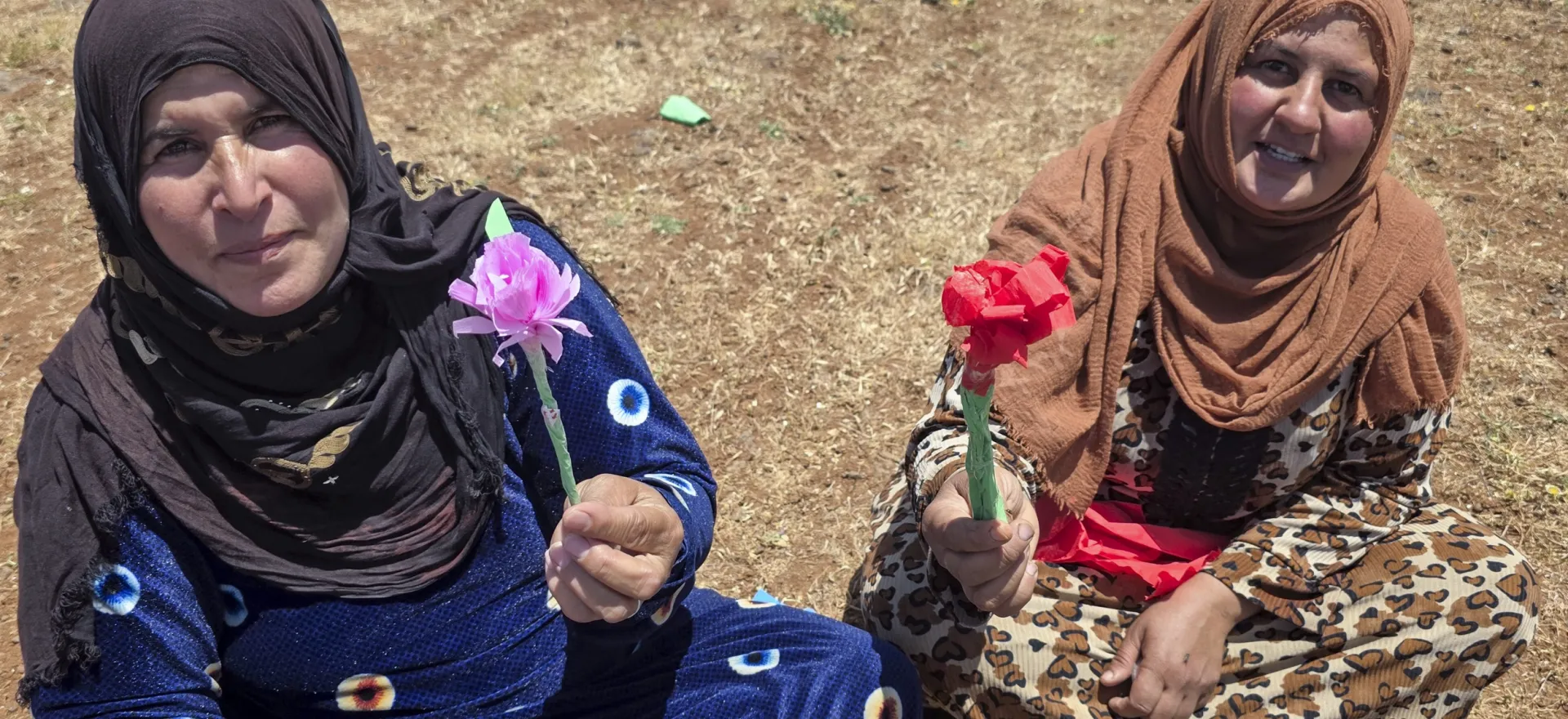 Two women sitting on the ground each hold a paper flower, the color of the dresses.