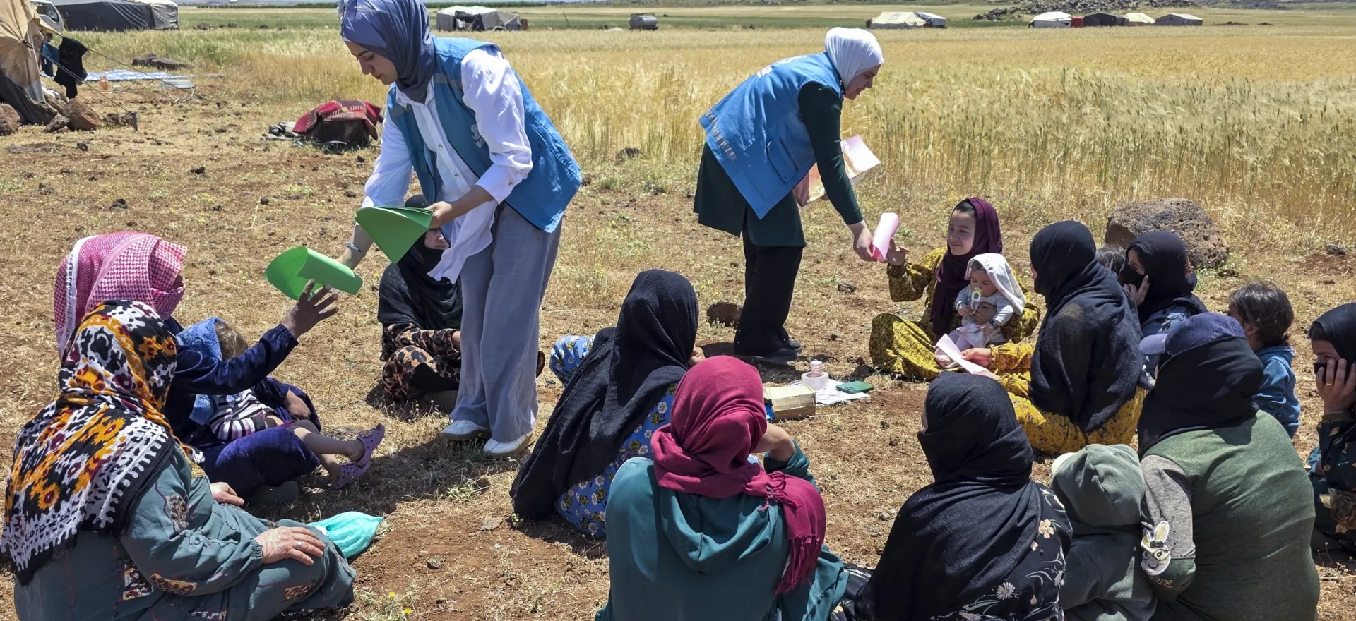 Two women wearing blue waistcoats hand out colored paper to women sitting on the ground in groups.