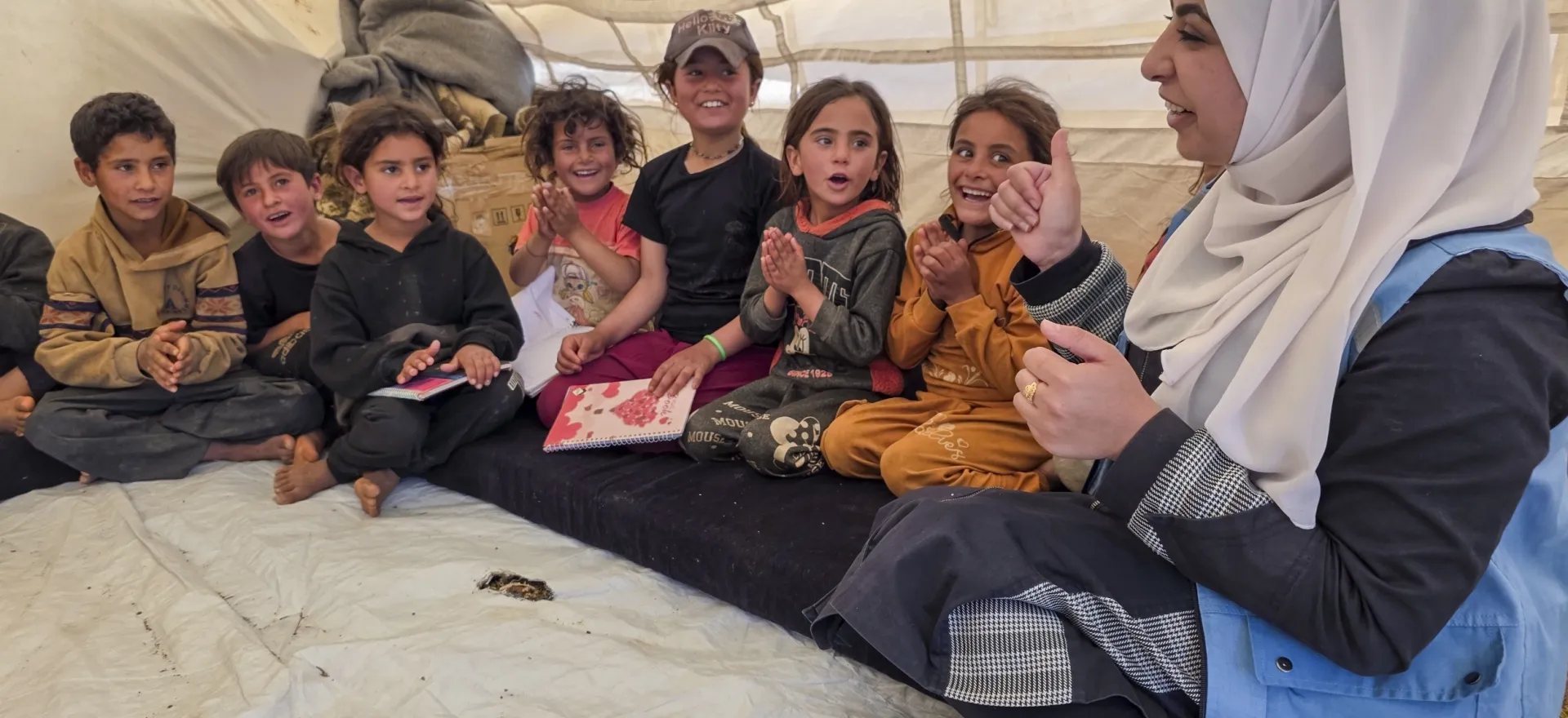 A group of children sit next to a female facilitator wearing a white headscarf inside a tent.