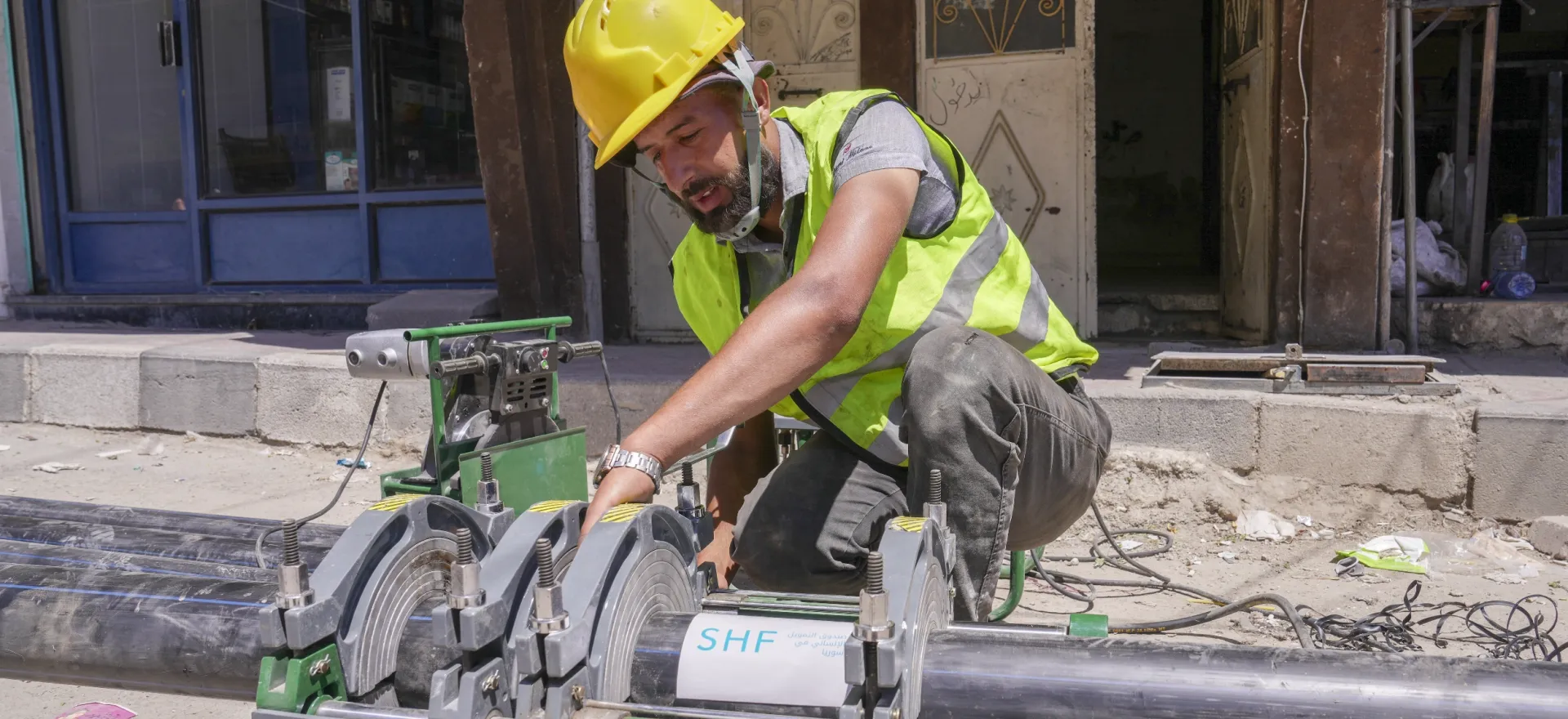 A worker, wearing a yellow reflective vest and a yellow hardhat, connects new water pipes before they are laid underground in Babila.