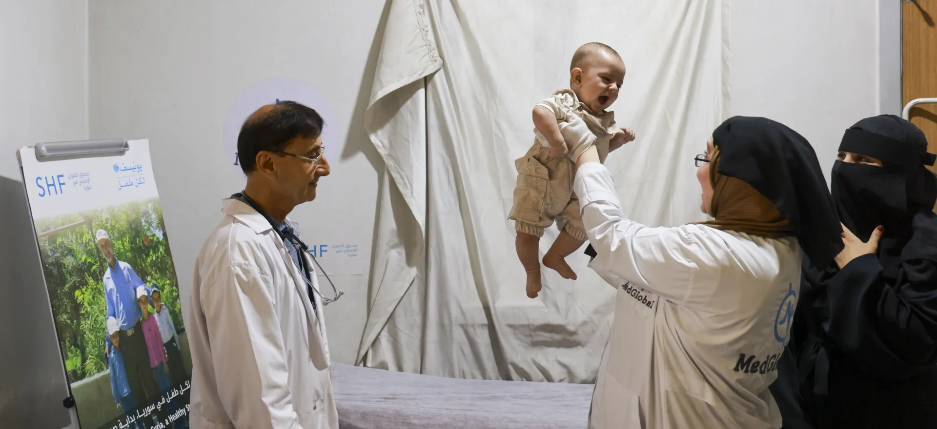 A woman wearing a white coat lifts a baby into the air.