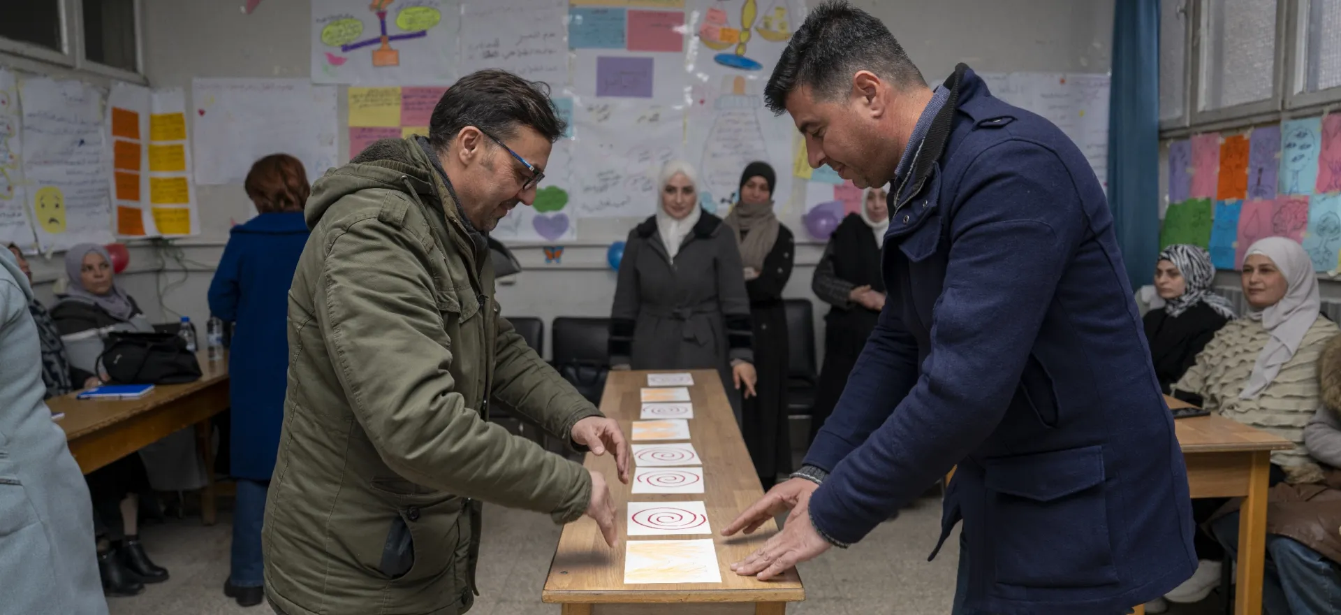 Two men standing on opposite sides of a long narrow table, concentrate on some cards that are laid out in front of them.