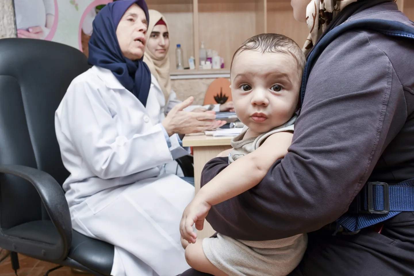 A baby looks at the camera while his mother speaks to a paediatrician