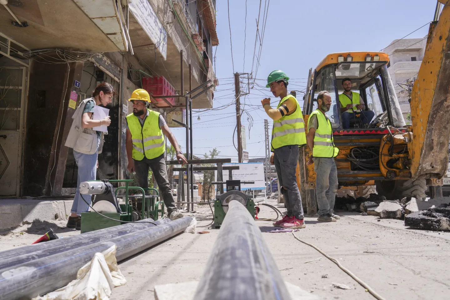 Heavy machinery works through a street in Babila, where UNICEF officer speaks to workers dressed in yellow reflective vests and hardhats, with long pipes lined up along the road.