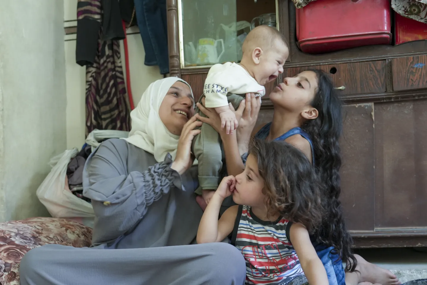 A girl with long black hair, sitting between her mother and younger sister, lifts a baby boy into the air.