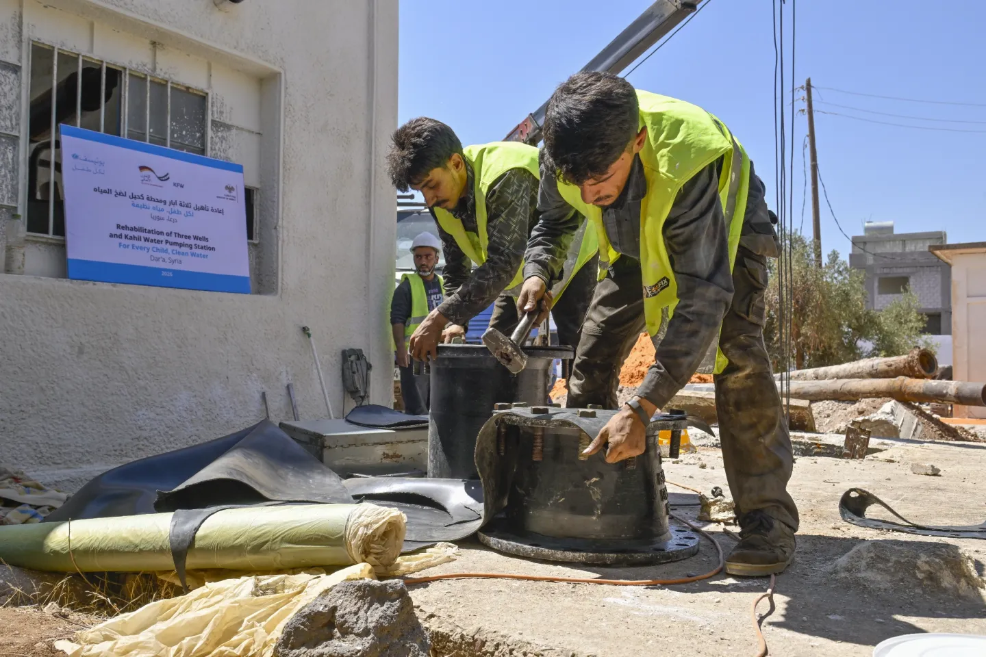 Two men wearing yellow reflective vests lift a heavy piece of equipment.