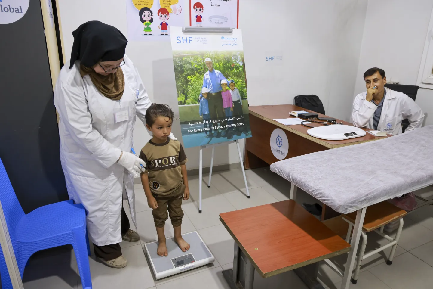 A woman with a black headscarf and white coat helps a little boy to stand on a scale, with a doctor sitting behind a desk looking on.