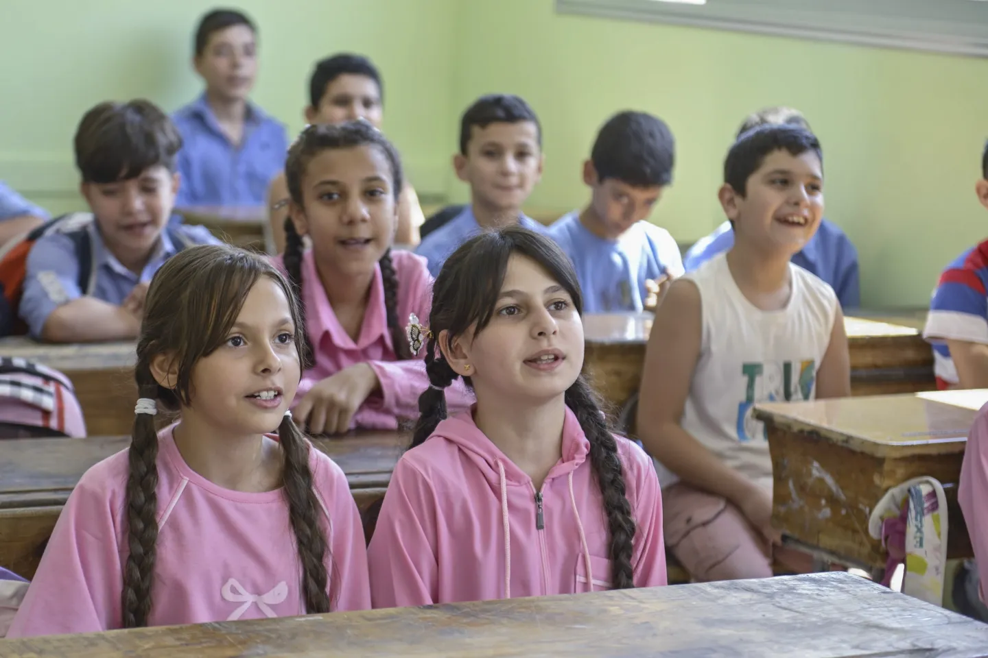 Two girls wearing pink school uniforms sit together in class with boys in blue and more girls in pink school uniforms.