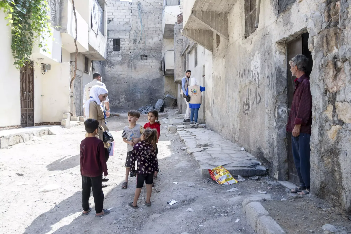 A female volunteer speaks to a group of young children in the street, while others are speaking to parents in doorways further down.
