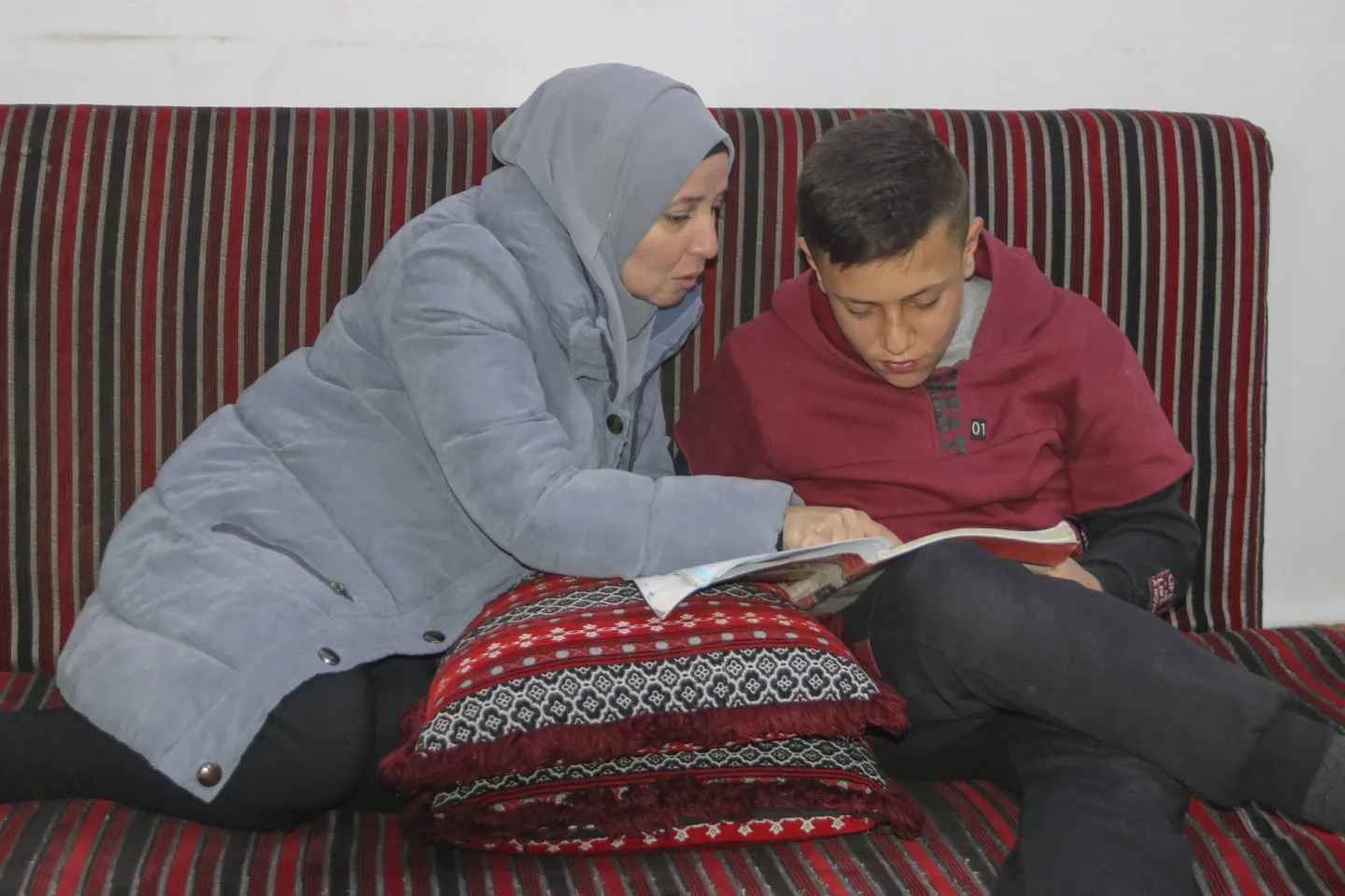 A mother leans over to look at the homework her son is doing in a book he his holding on his lap.