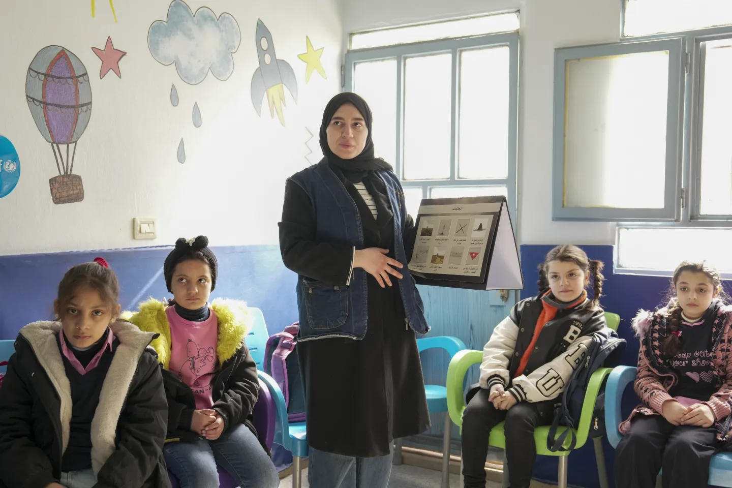 A woman shows a poster with pictures on it to a group of children in a classroom.