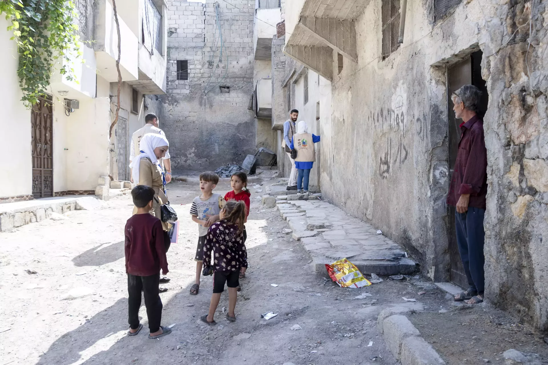 A female volunteer speaks to a group of young children in the street, while others are speaking to parents in doorways further down.