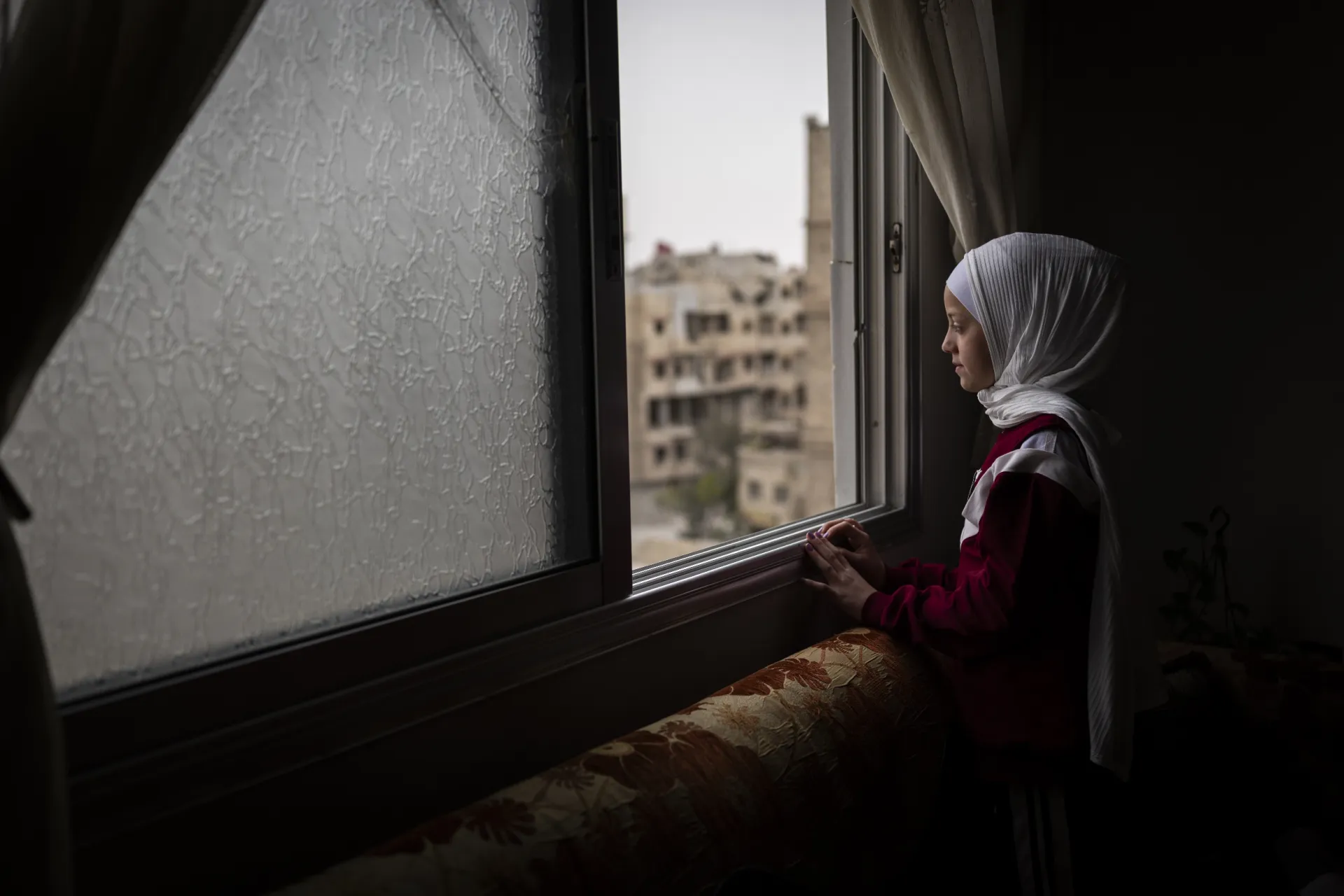 A girl looks out a window with damaged buildings visible outside