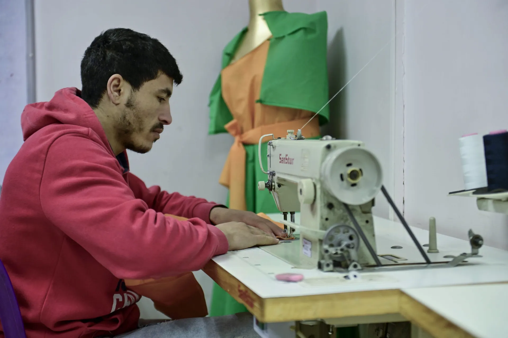 A young man wearing a red hoodie, works behind a sewing machine.