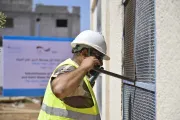 A man wearing a white hardhat and yellow reflective vest uses a large tool to install a metal mesh.