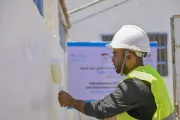 A man wearing a white hard-hat and a yellow reflective vest, paints a wall.