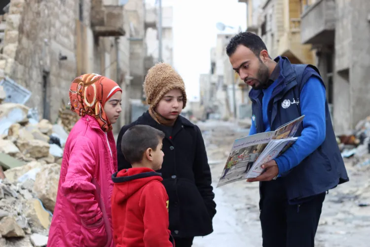 A volunteer show pictures to three young children in a street with rubble and war damaged buildings.