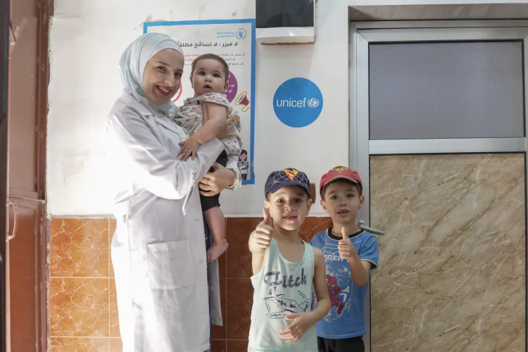 A female physician, wearing a white coat and headscarf, holds a baby girl in her arms with the baby's two little boys standing next to her.