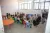 Young children sit on colourful little chairs in two rows, listening to a female who sits at one end of the two rows.