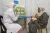 A woman wearing all white clothing, points at a poster displaying pictures of breastfeeding, with a mother holding her baby looking and listening.