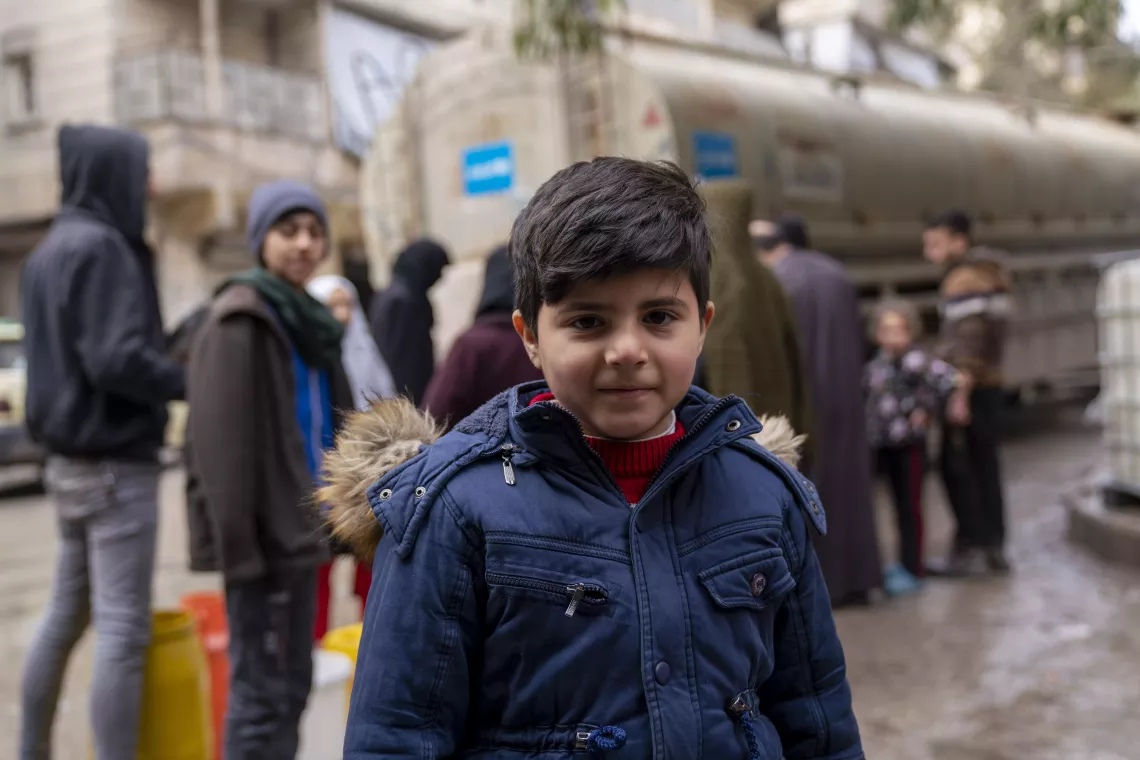 A boy stands in front of a water truck with a UNICEF logo on it and a crowd of people waiting to collect water.
