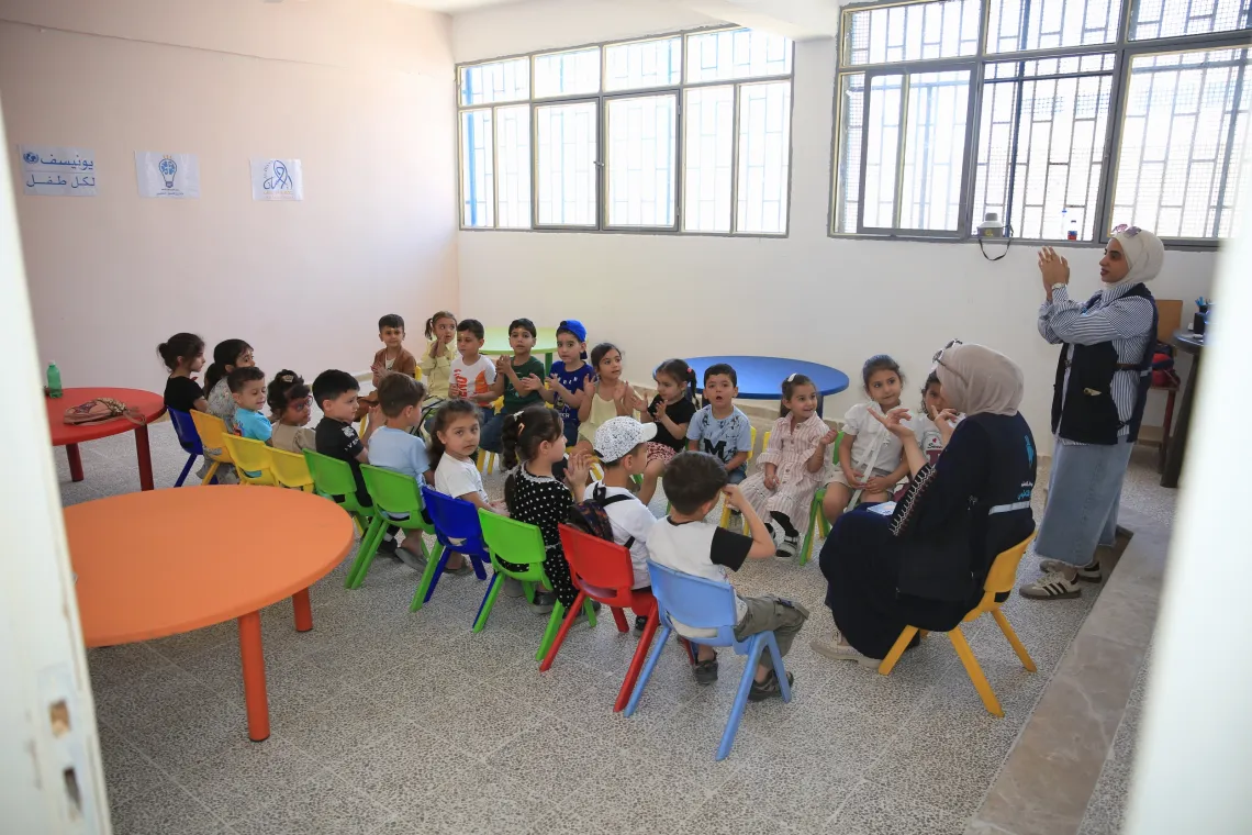 Young children sit on colourful little chairs in two rows, listening to a female who sits at one end of the two rows.