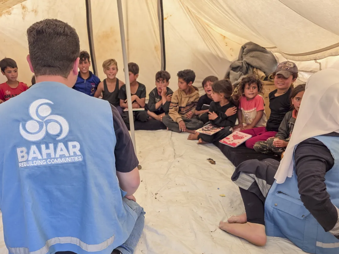 Facilitators wearing blue waistcoats sit with children in a circle on the floor of a tent.