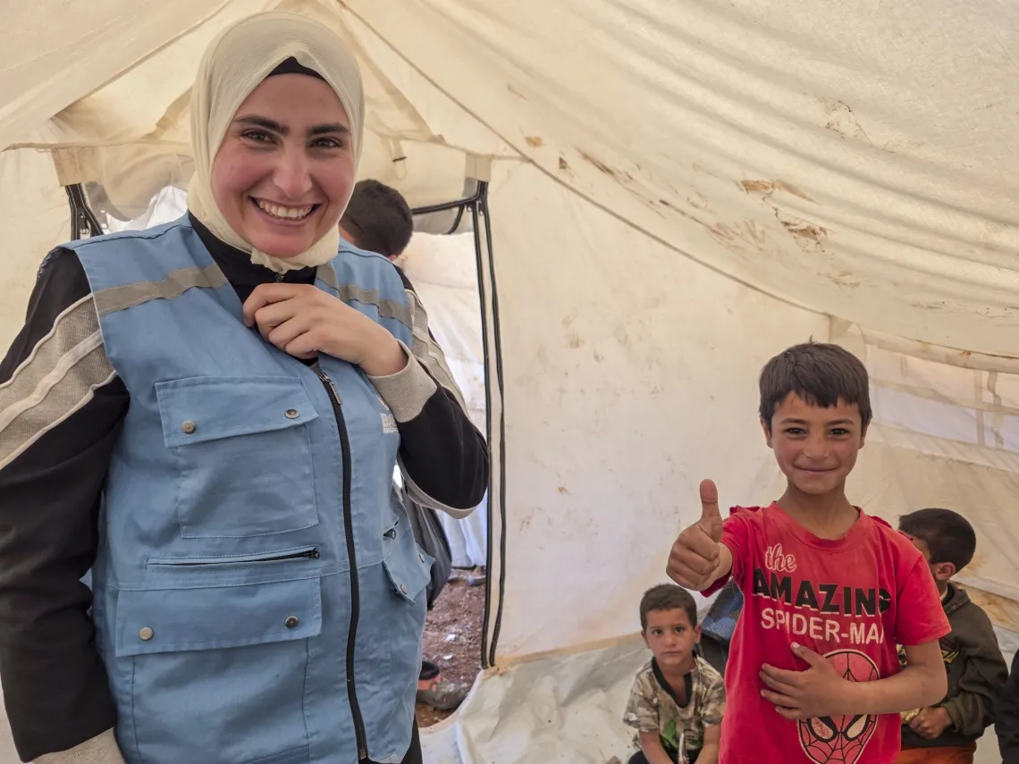 A little boy wearing a red t-shirt gives a thumbs up, with a smiling facilitator wearing a blue waistcoat and white headscarf standing to his side. 
