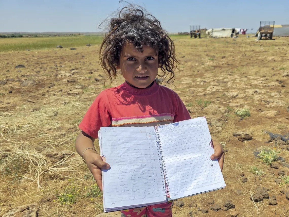 A little girl wearing a red t-shirt holds her practice book open in front of her.