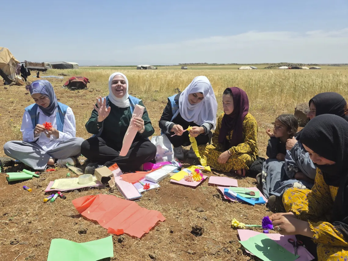 Women sit next to each other on the ground with colored papers spread out in front of each of them. of 