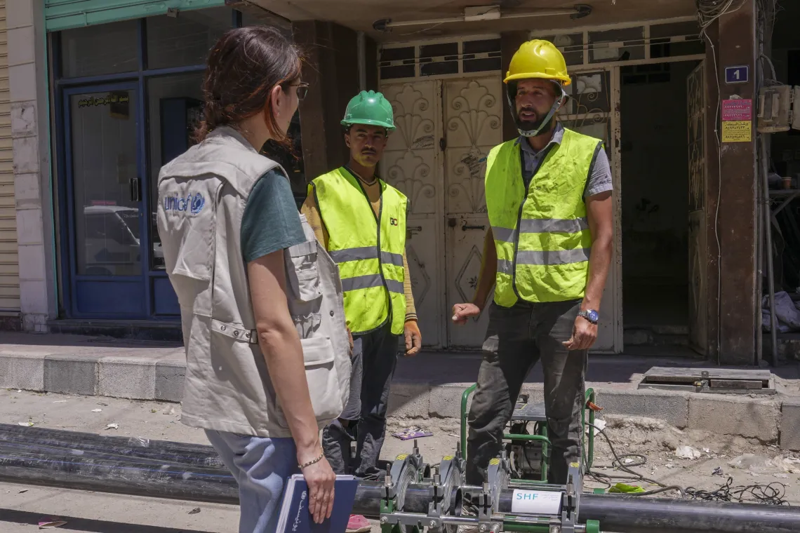 A woman wearing a UNICEF waistcoat, chats with two workers wearing yellow reflective jackets and yellow and green hardhats.