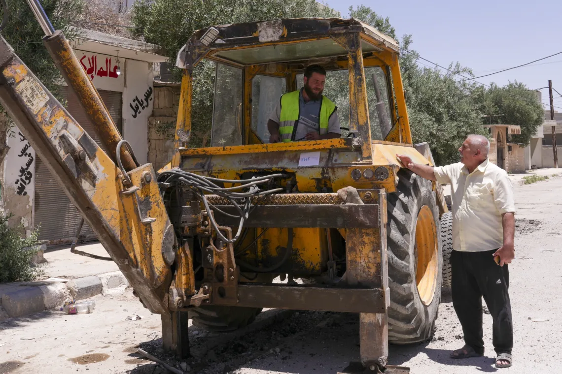 A man wearing a yellow shirt, chats with a worker who is sitting in large yellow excavator.