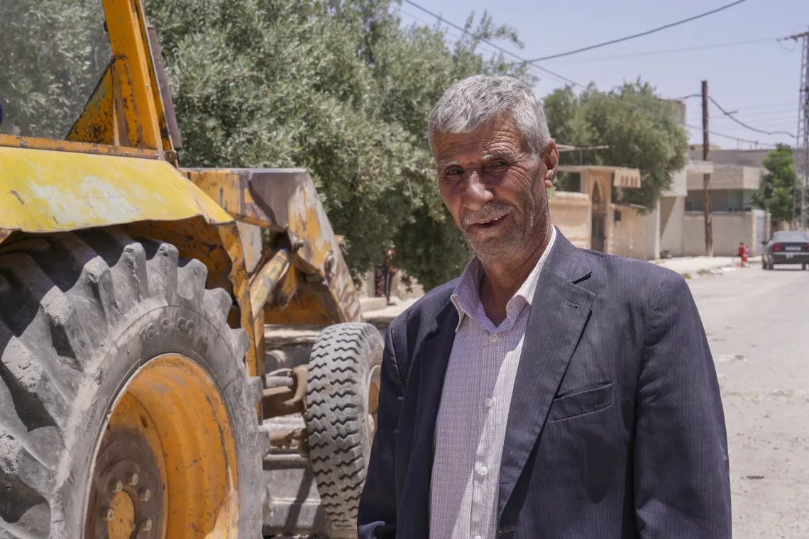 A man wearing a shirt and suit jacket stands next to large yellow excavator.
