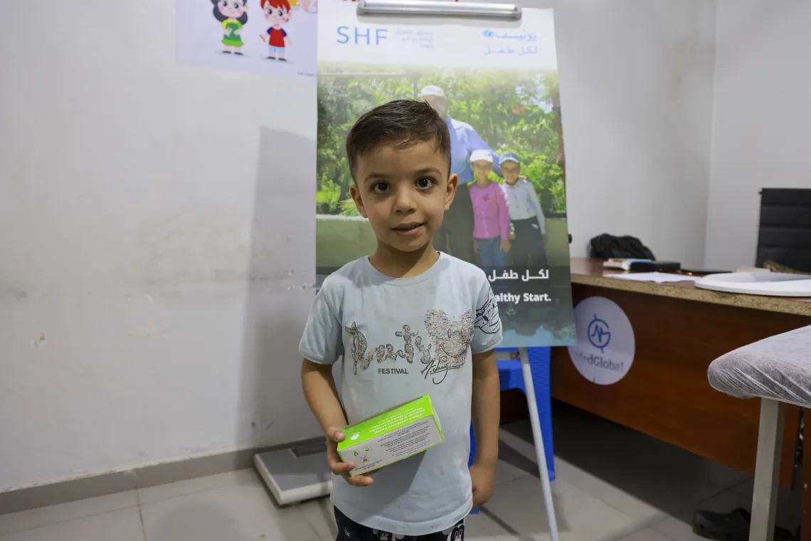 A young boy holding a box in his right hand stands in health centre.