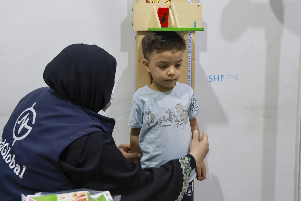 A boy stands with his back against a wall with a device resting on his head to measure his height.