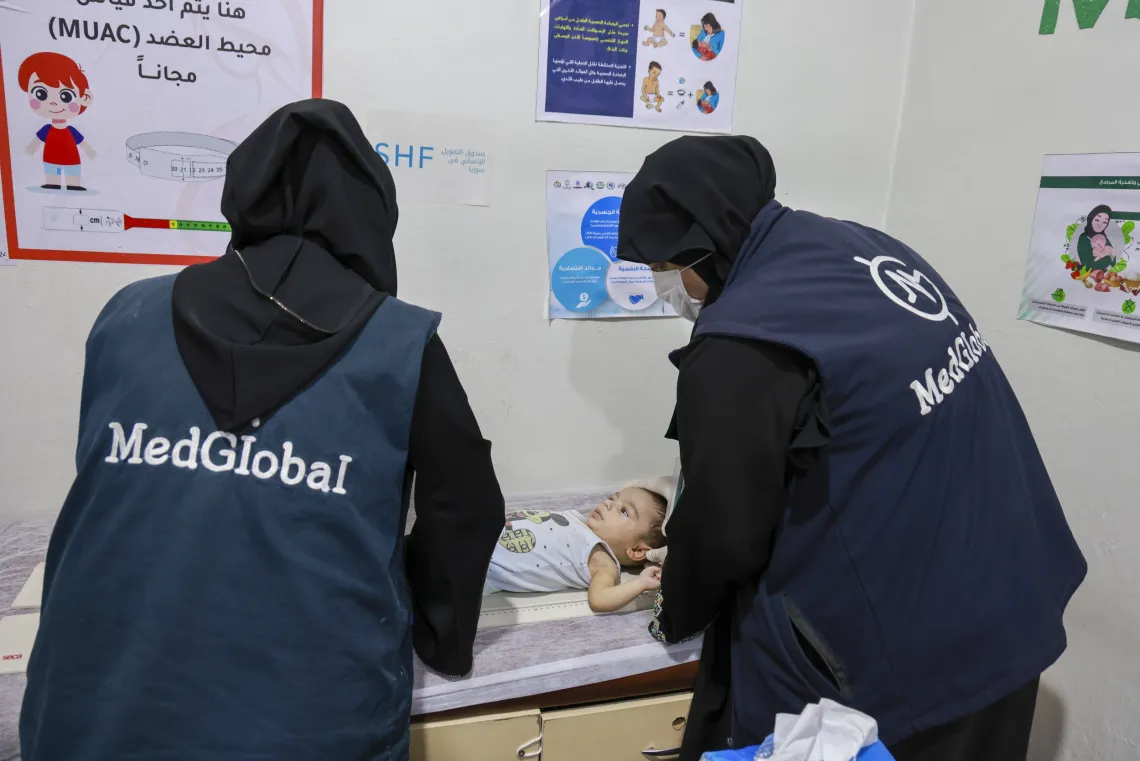 A baby boy is lying on his back on an examination table, while two health worker measures his height.
