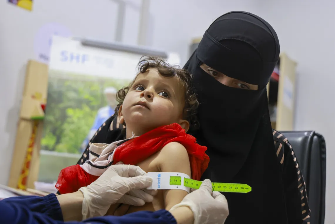 A girl wearing a red top looks up from where sits on her mother's lap, while her mid-upper arm circumference is measured.