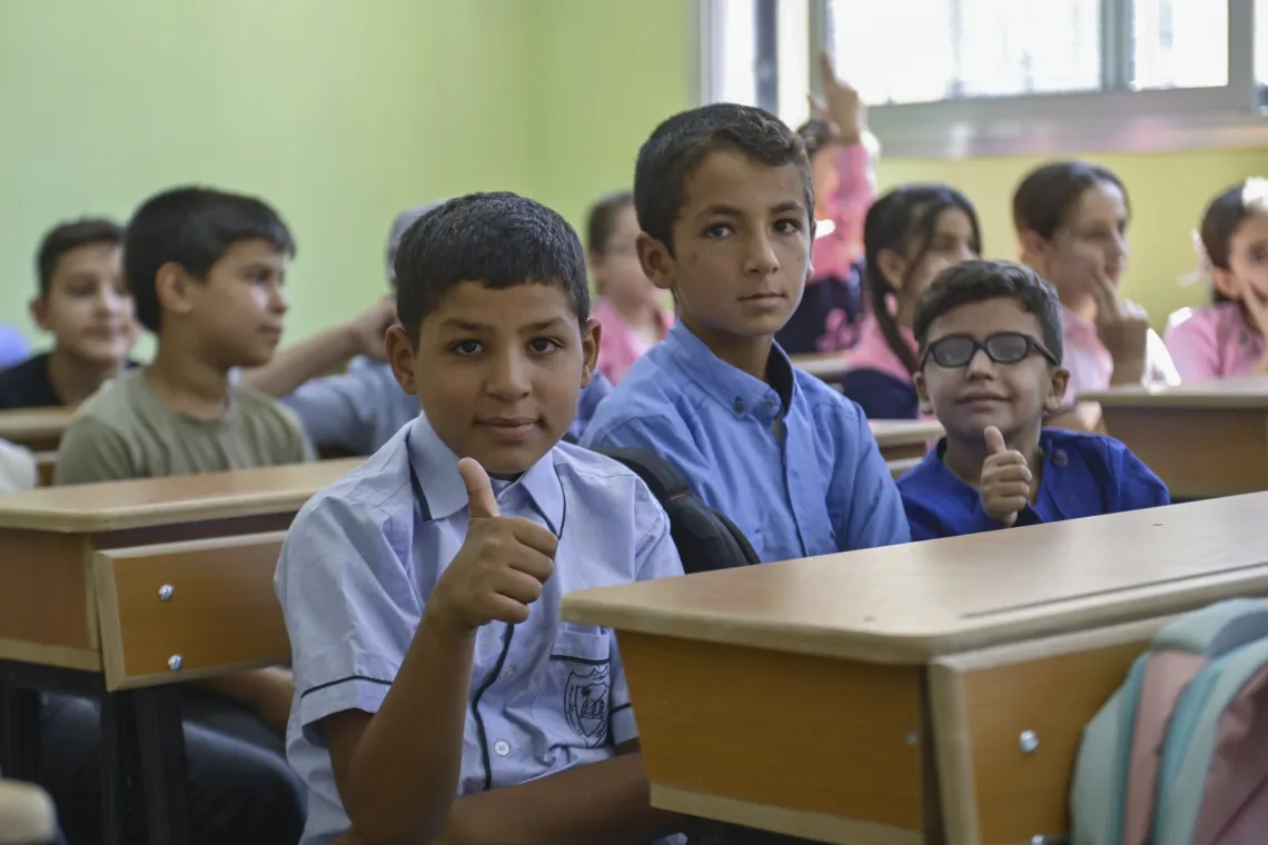 A boy sits next to two others at a desk in a class. They all wear blue school uniforms.