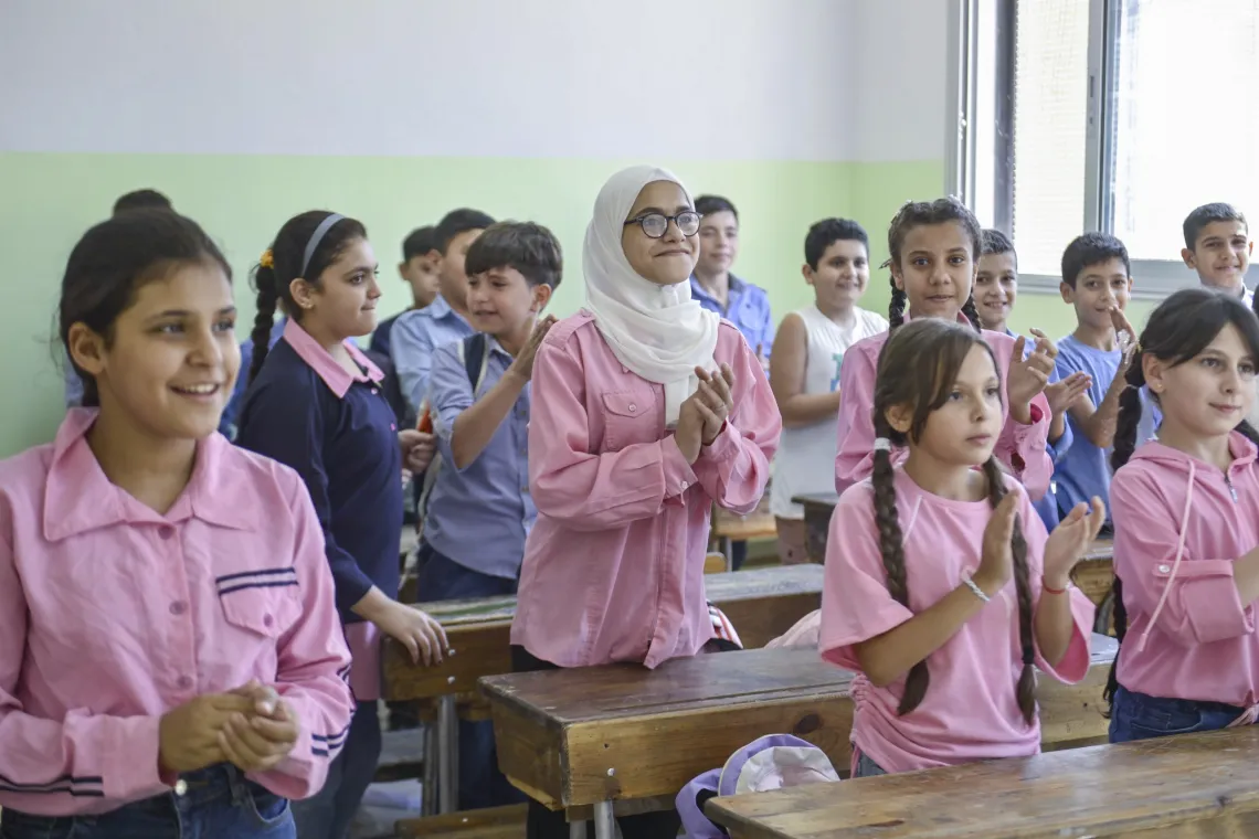 Children stand behind their desks in class. Girls wearing pink uniforms and the boys blue.