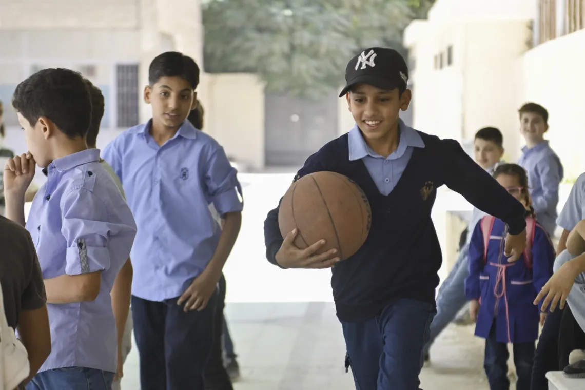 A boy with a basketball under his arm runs past his classmates outside class in a school courtyard.