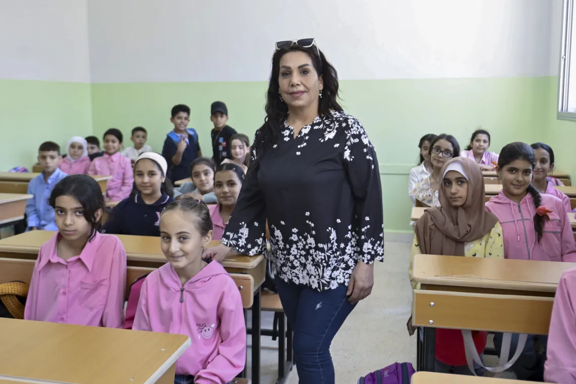 A female teacher stands behind in the isle of a classroom filled with girls and boys wearing pink and blue school uniforms.
