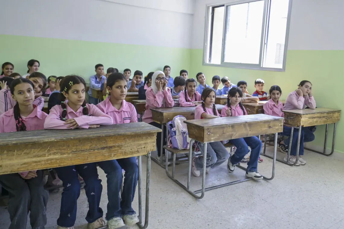 Girls wearing pink and boys blue school uniforms sit at desks in a classroom.