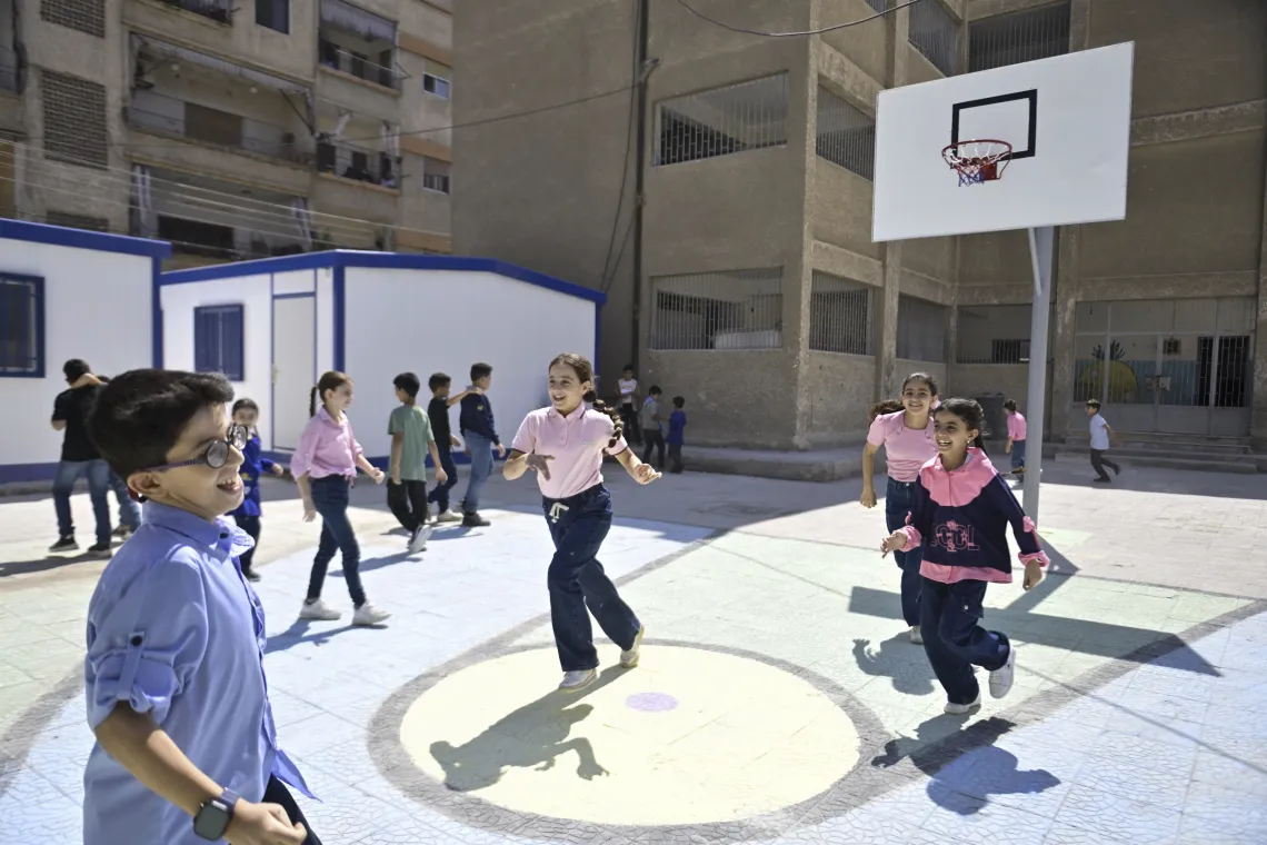 Children wearing school uniforms run and play in a courtyard on a basketball court.