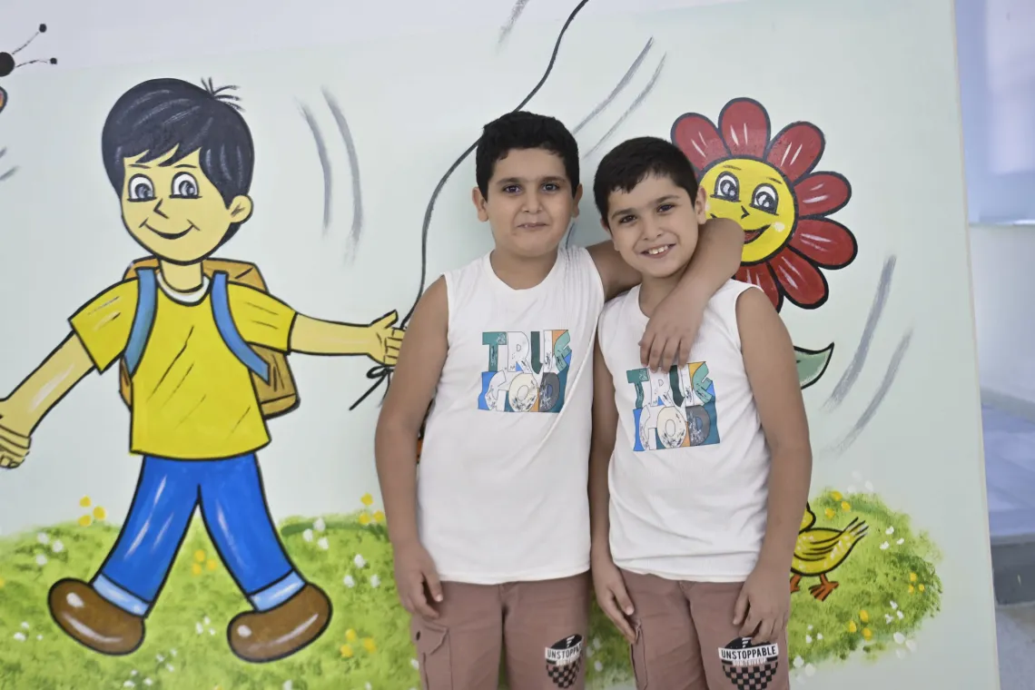 Two similar looking boys with one arm around the others shoulder pose for a photo in front of a colourful mural.