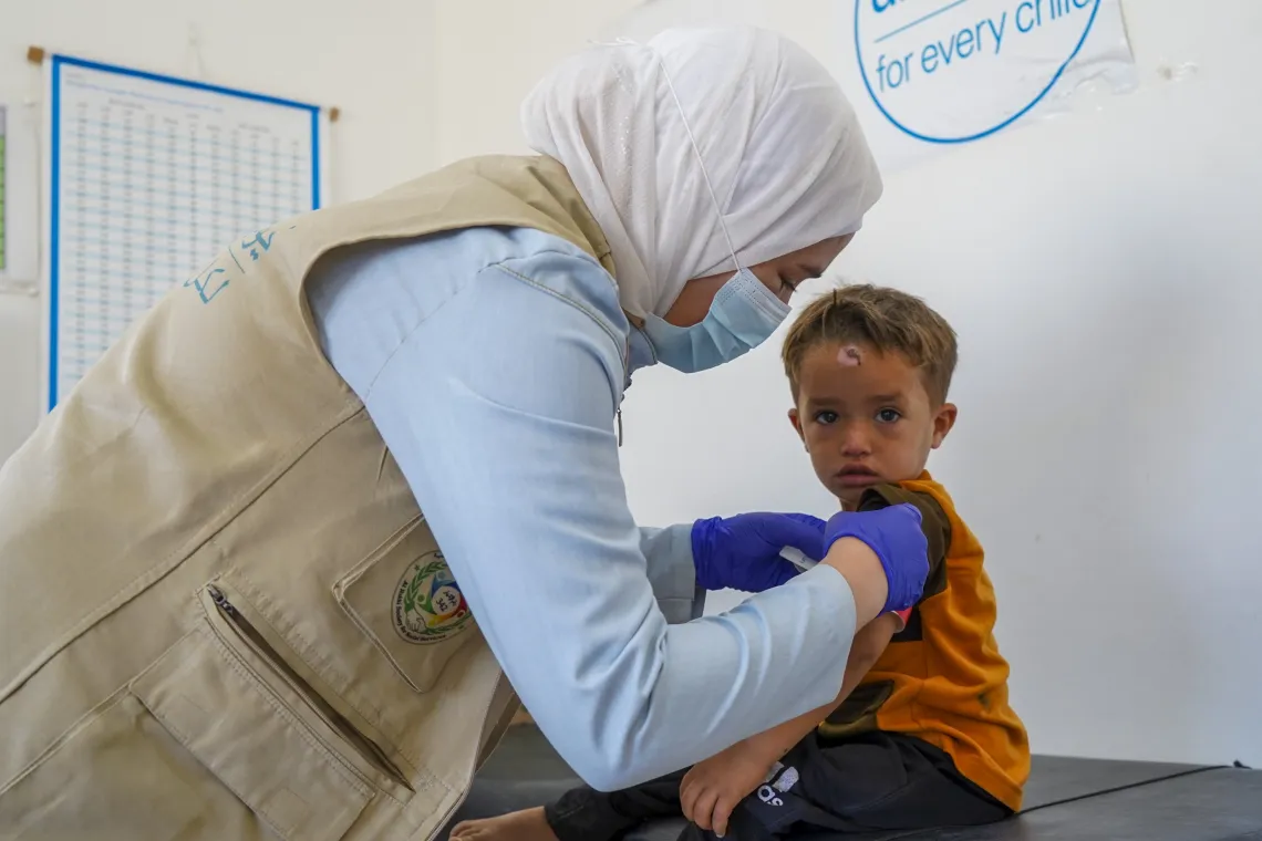 A little boy looks up while a female wearing a white headscarf measures his mid-upper arm circumference.