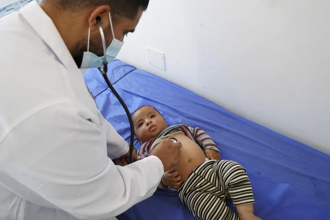 A doctor with a stethoscope bends over a small child who lying on his back on a blue examination table.
