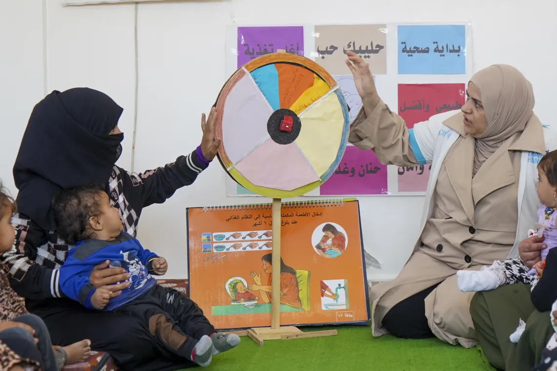 A woman sitting on a carpet, holding a colourful cutout of a pie chart with a poster underneath it. Another woman holds it up from the other side.