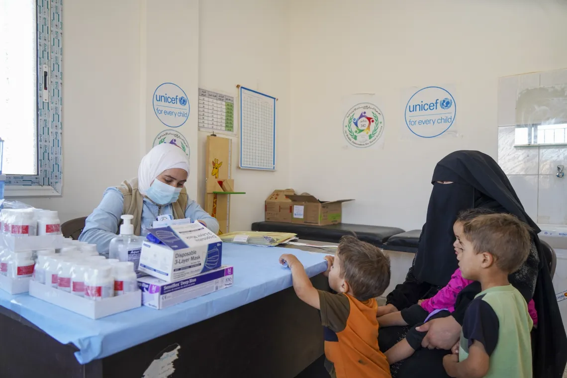 A nurse sits behind a blue desk with medical supplies stacked to the one side. Opposite her is a seated woman with three children, one on her lap and two standing next to her.
