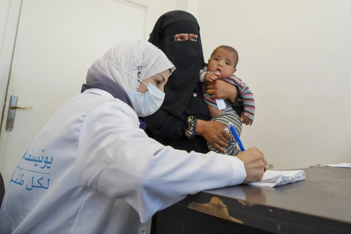 A woman holding a baby on her him looks on while a woman in white coat makes some notes in a book.