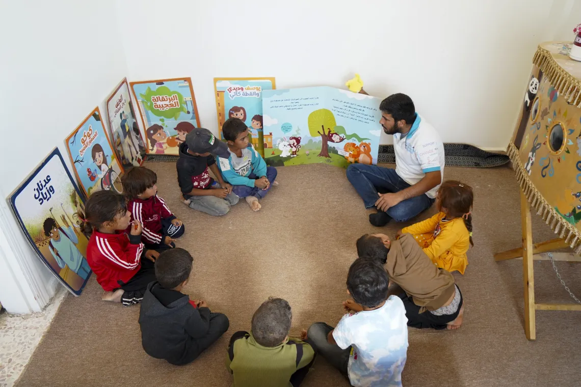 A young man sits crossed-legged with children on the floor, with colourful posters behind them.